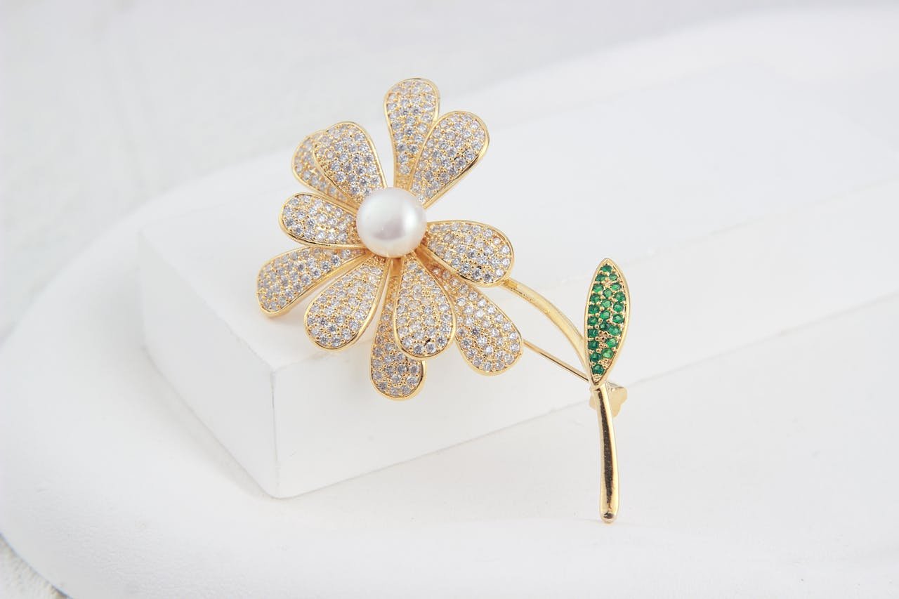 Close-up of a luxurious flower-shaped gold brooch adorned with jewels and a pearl centerpiece on a white background.