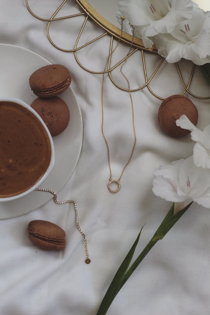 A flat lay of macarons, coffee, gold jewelry, and white flowers on a white cloth.